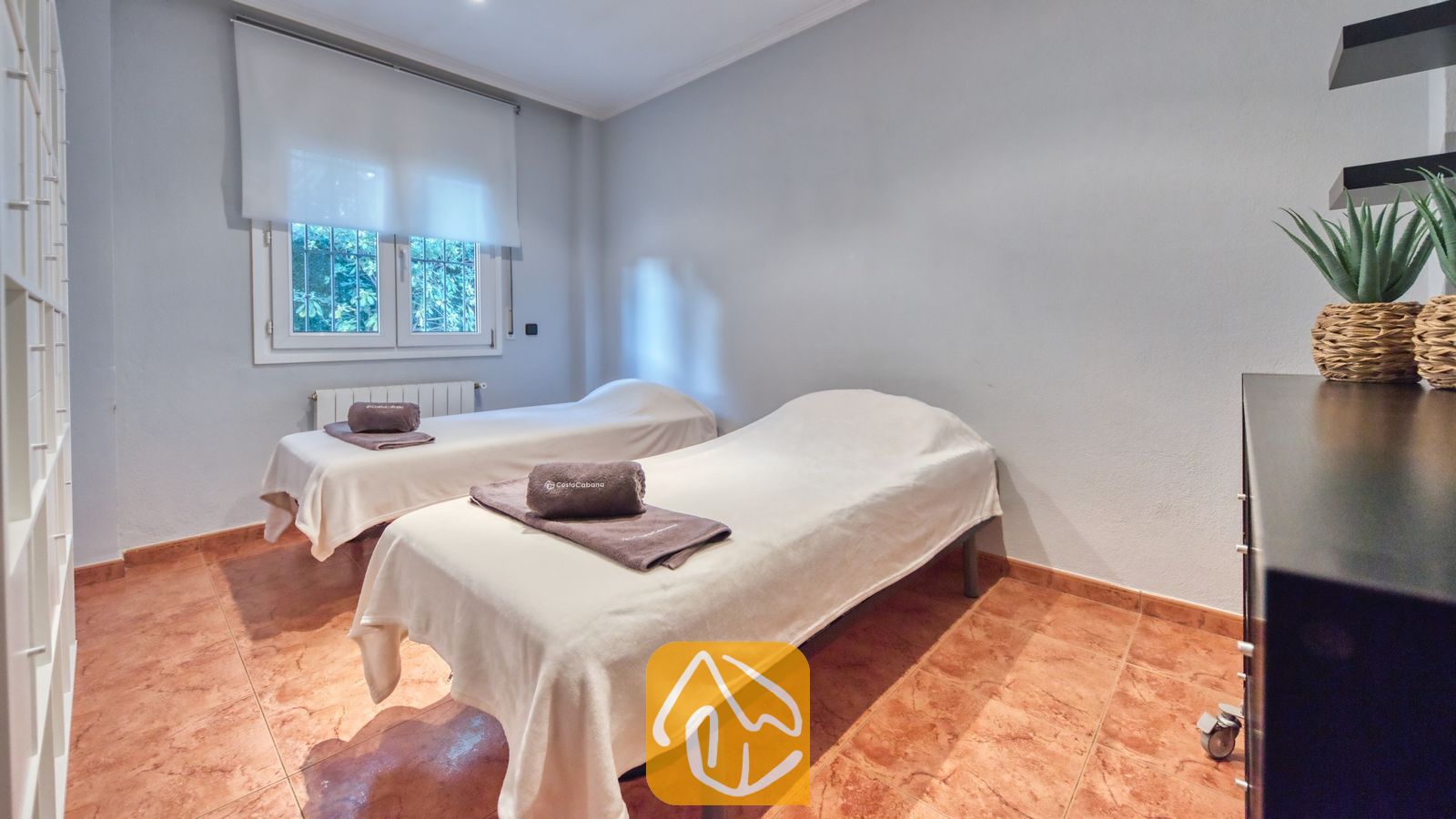 Vakantievilla's Costa Brava Spanje - Villa Summertime - Massagekamer met twee behandelbedden voorzien van witte lakens, elk met een opgevouwen grijze handdoek erop, tegelvloer en een raam met een wit rolgordijn.
