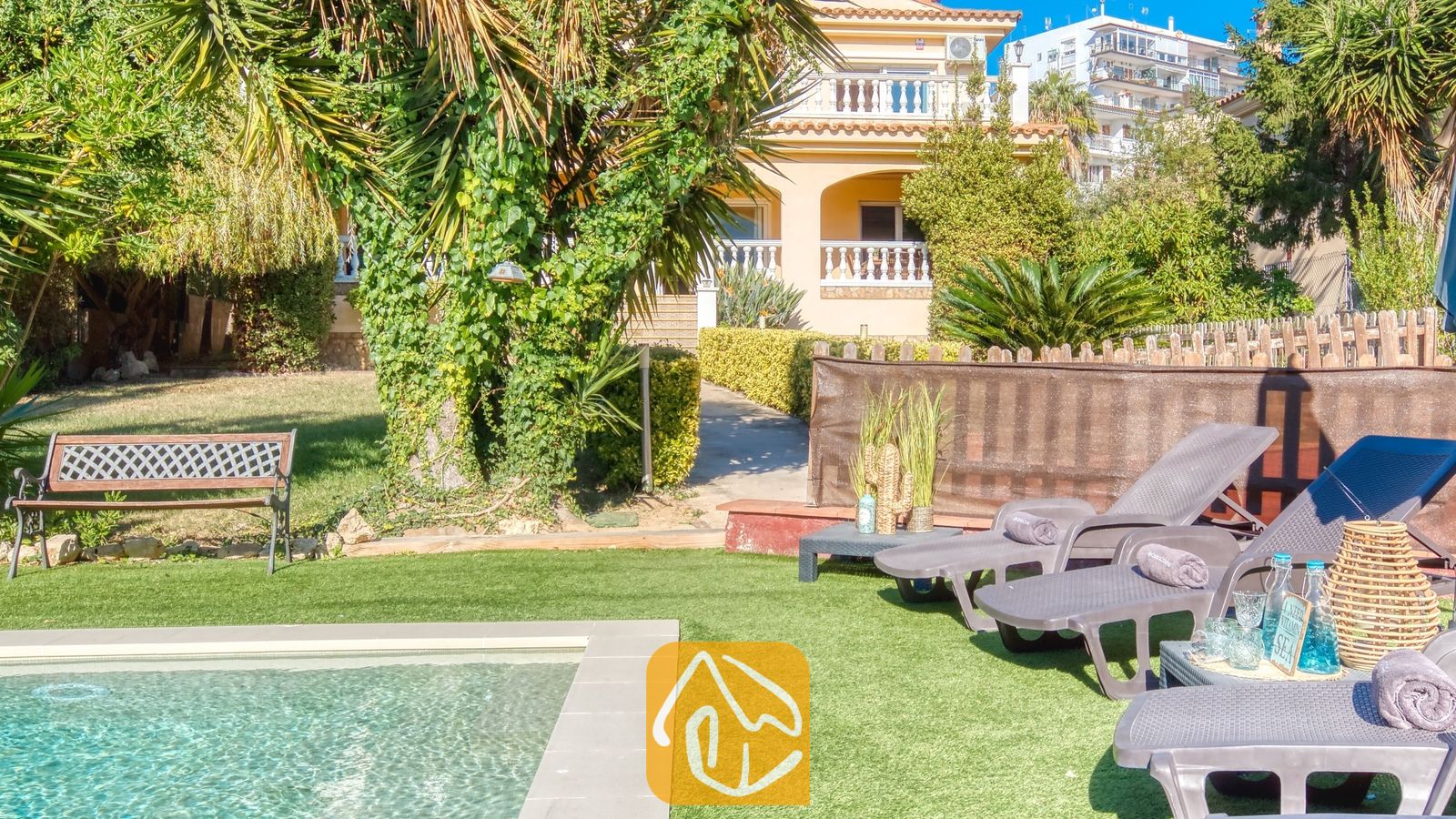 Vakantievilla's Costa Brava Spanje - Villa Summertime - Zonnige tuin bij Villa Summertime met grijze ligstoelen naast een zwembad, kunstgras en een geel gebouw van meerdere verdiepingen op de achtergrond.