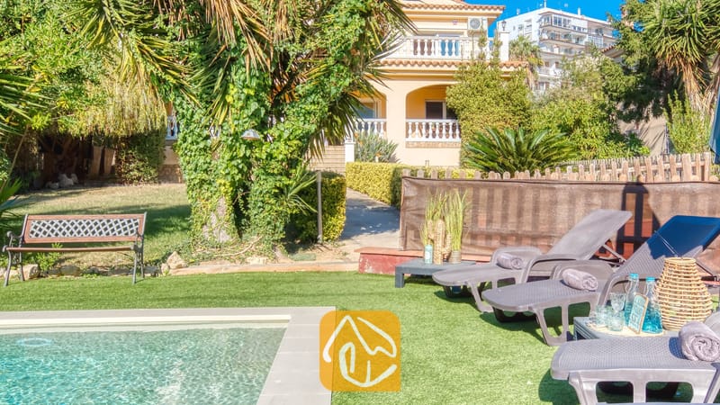 Vakantievilla's Costa Brava Spanje - Villa Summertime - Zonnige tuin bij Villa Summertime met grijze ligstoelen naast een zwembad, kunstgras en een geel gebouw van meerdere verdiepingen op de achtergrond.