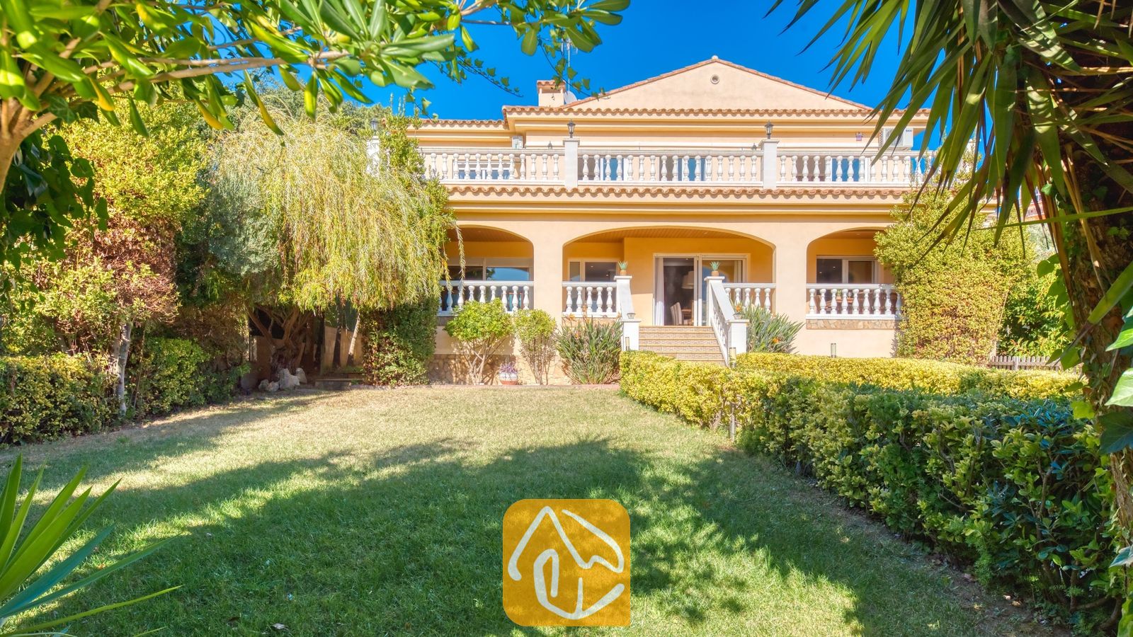 Vakantievilla's Costa Brava Spanje - Villa Summertime - Buitengevel van Villa Summertime met een beige gebouw van twee verdiepingen, een terras met een witte balustrade, een groen gazon en omliggende tuinhagen onder een blauwe lucht.