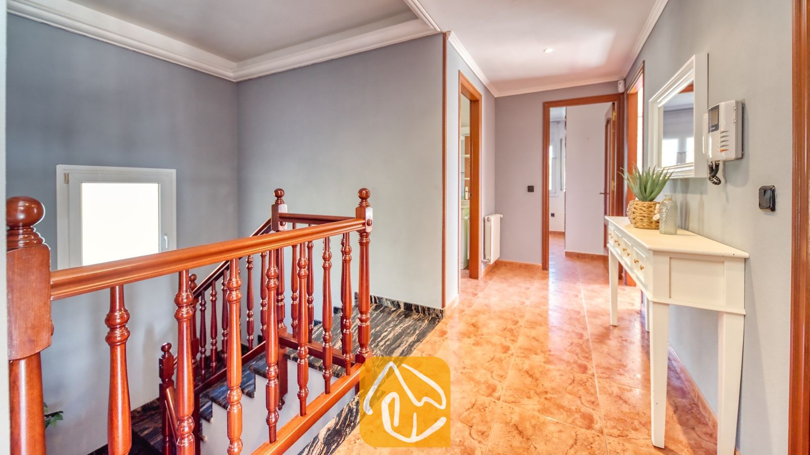 Vakantievilla's Costa Brava Spanje - Villa Summertime - Houten trapeluning die naar beneden leidt langs een hal met tegelvloeren, een witte sidetable en grijze muren in Villa Summertime.