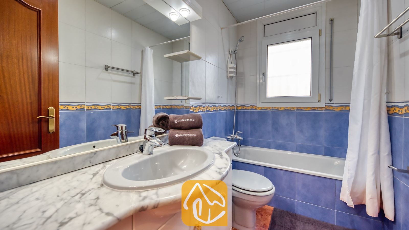 Vakantievilla's Costa Brava Spanje - Villa Summertime - Badkamer voorzien van een wastafel met marmeren blad, blauw-witte tegelmuren, een ligbad met douche en opgerolde bruine handdoeken.
