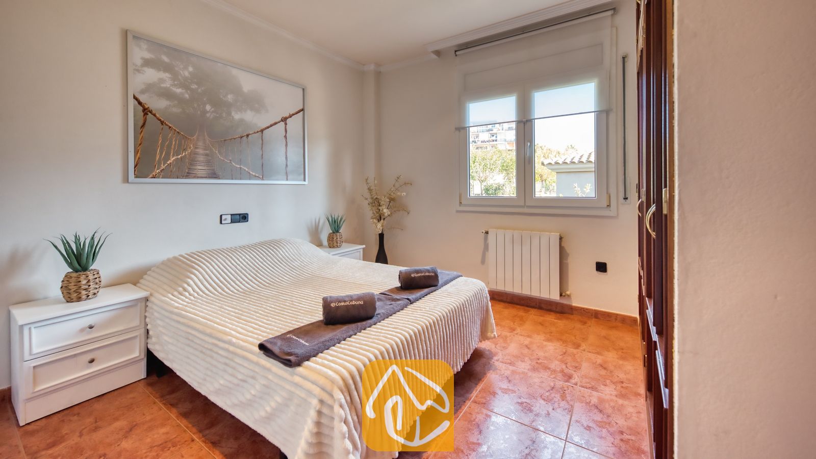 Vakantievilla's Costa Brava Spanje - Villa Summertime - Slaapkamer met een tweepersoonsbed, witte sprei met textuur, opgevouwen bruine handdoeken, twee witte nachtkastjes met in pot geplante planten en een grote ingelijste natuurfoto aan de muur.