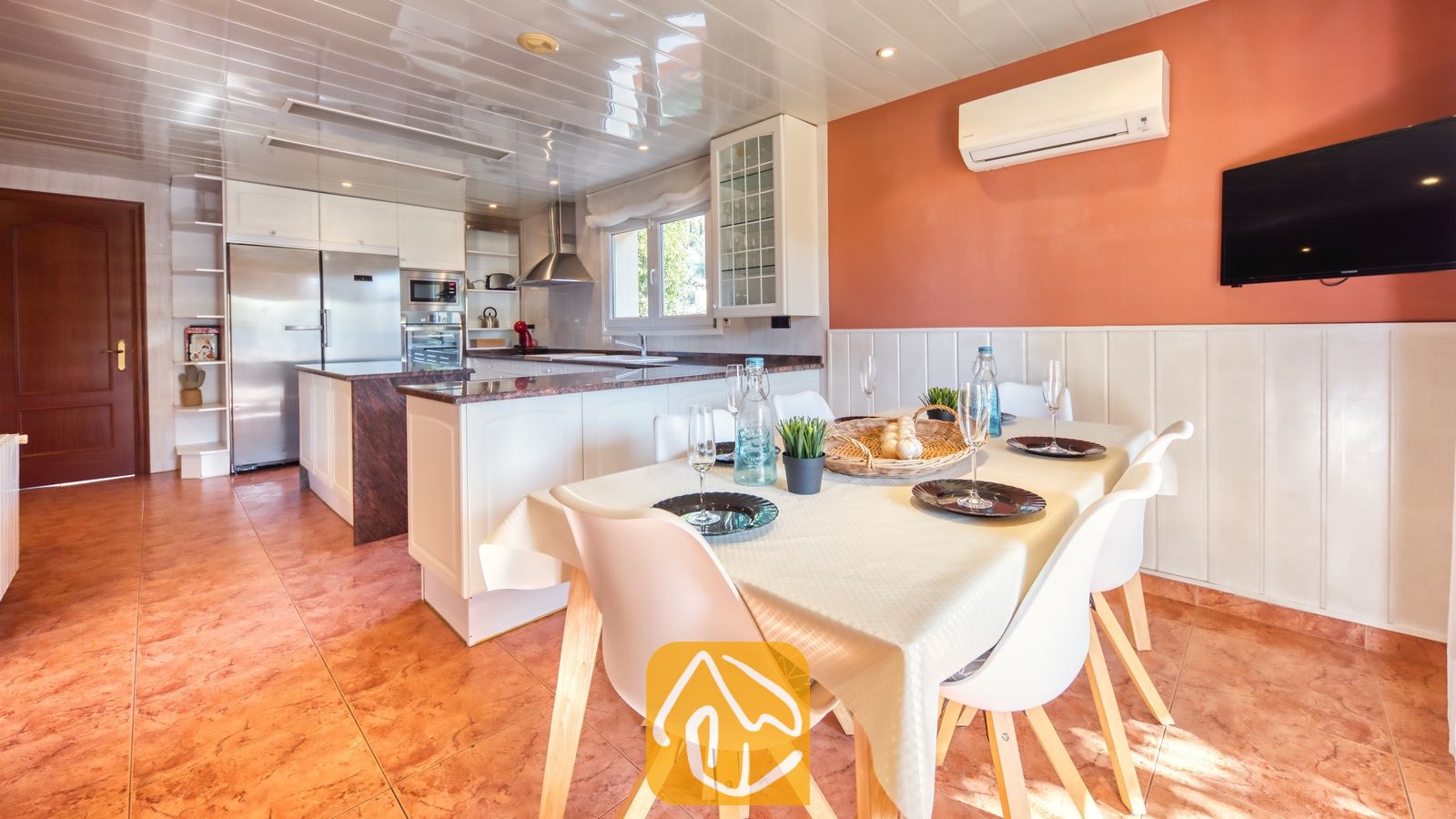 Vakantievilla's Costa Brava Spanje - Villa Summertime - Eethoek met een met wit linnen gedekte tafel en witte stoelen naast een open keuken bij Villa Summertime, voorzien van terracotta tegelvloeren en oranje muren.