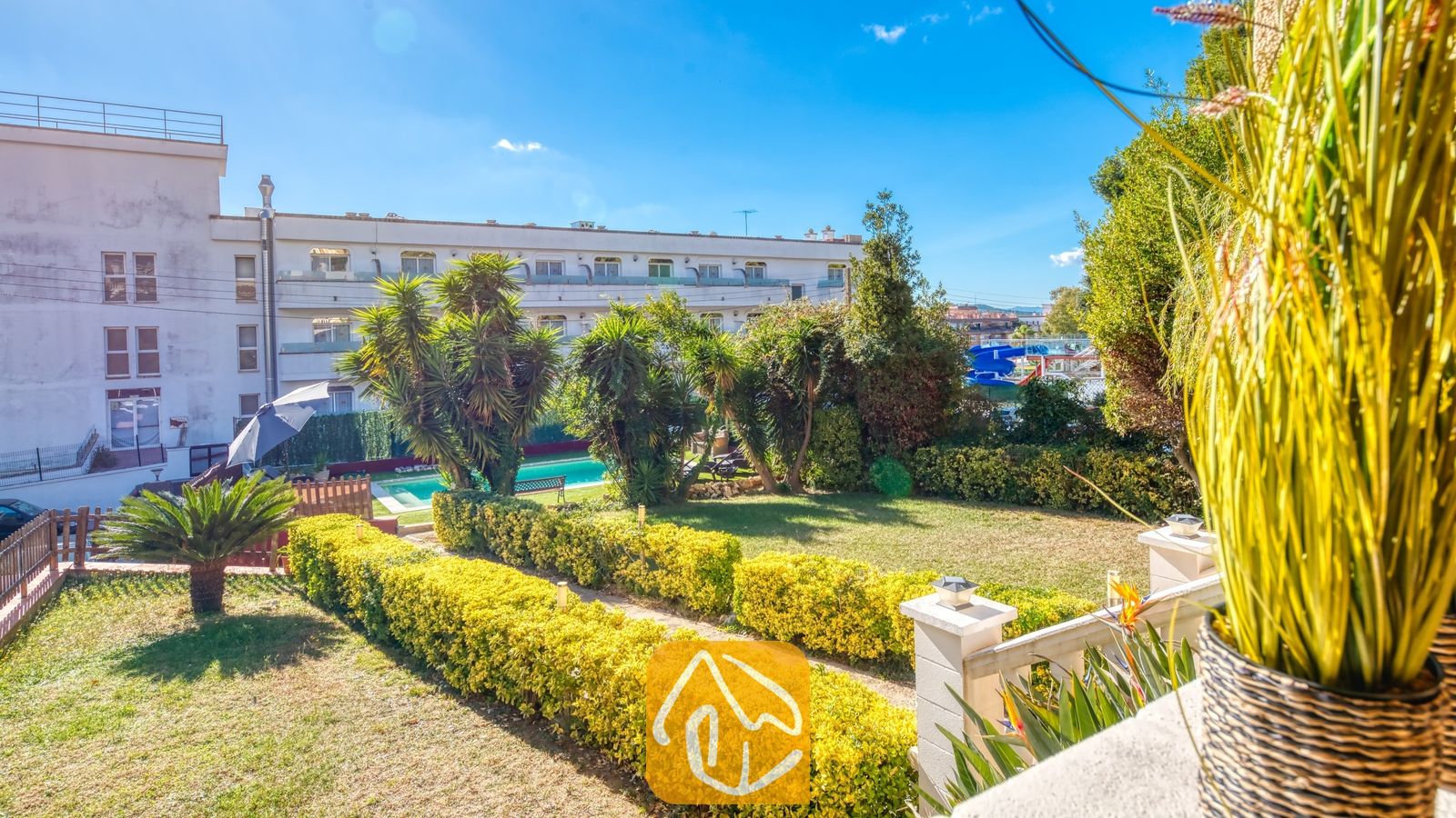 Vakantievilla's Costa Brava Spanje - Villa Summertime - Zonnig tuinzicht vanuit Villa Summertime met strak gesnoeide gele heggen, een groen gazon, palmbomen en een zwembad op de achtergrond onder een blauwe hemel.