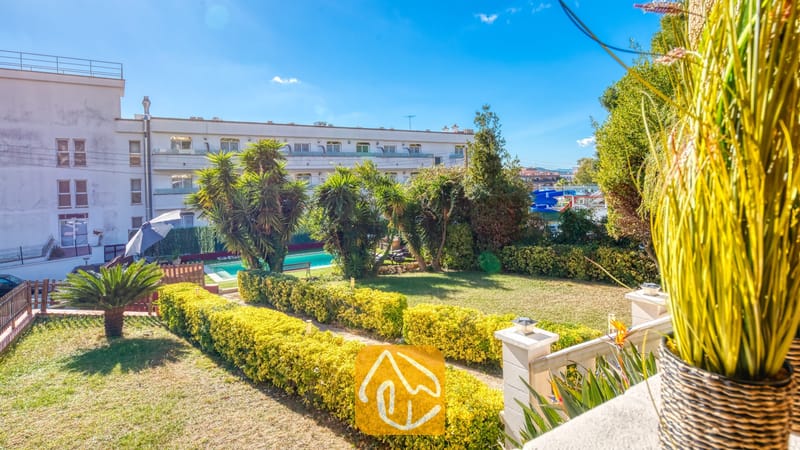 Vakantievilla's Costa Brava Spanje - Villa Summertime - Zonnig tuinzicht vanuit Villa Summertime met strak gesnoeide gele heggen, een groen gazon, palmbomen en een zwembad op de achtergrond onder een blauwe hemel.