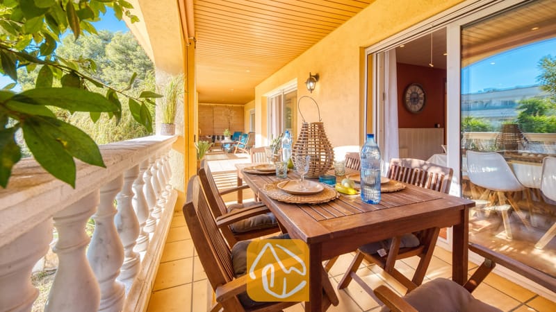 Vakantievilla's Costa Brava Spanje - Villa Summertime - Overdekt terras bij Villa Summertime, voorzien van een houten eettafel gedekt met geweven placemats, glaswerk en mineraalwater, omlijst door witte balustrade-hekwerken.