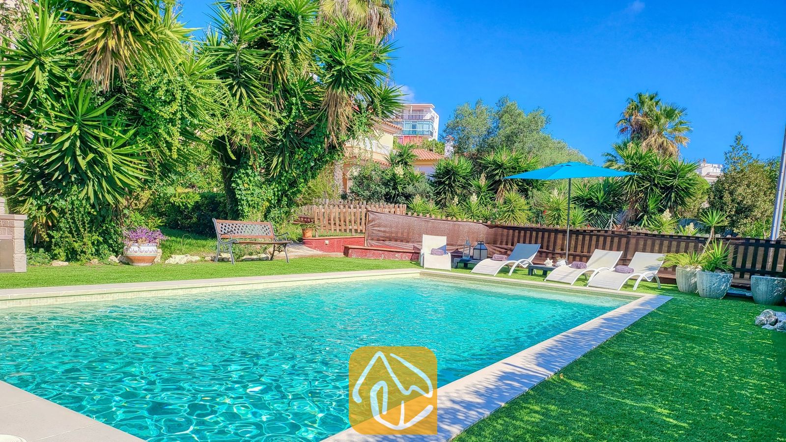 Vakantievilla's Costa Brava Spanje - Villa Summertime - Buitenzwembad bij Villa Summertime, omringd door een groen gazon, ligstoelen, een blauwe parasol en tropische palmbomen onder een helderblauwe hemel