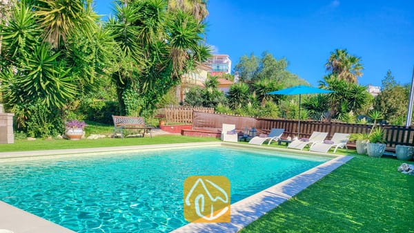 Vakantievilla's Costa Brava Spanje - Villa Summertime - Buitenzwembad bij Villa Summertime, omringd door een groen gazon, ligstoelen, een blauwe parasol en tropische palmbomen onder een helderblauwe hemel