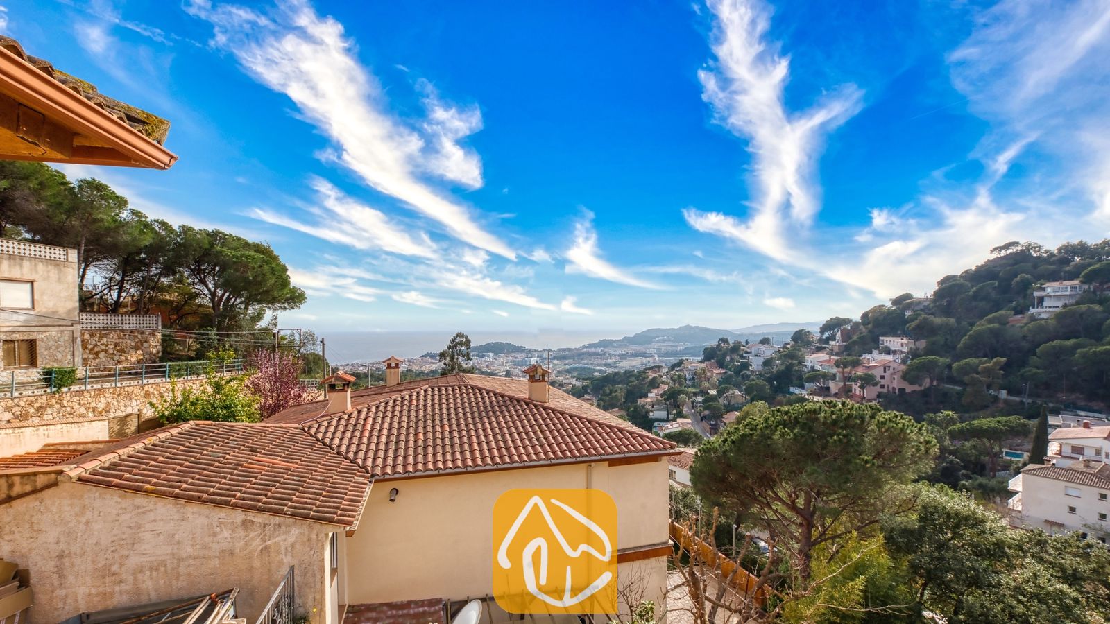 Vakantievilla's Costa Brava Spanje - Villa Marysol - Verheven uitzicht vanuit Villa Marysol met terracotta pannendaken, omringende groene heuvels en de verre zee onder een helderblauwe hemel met sluierwolken.