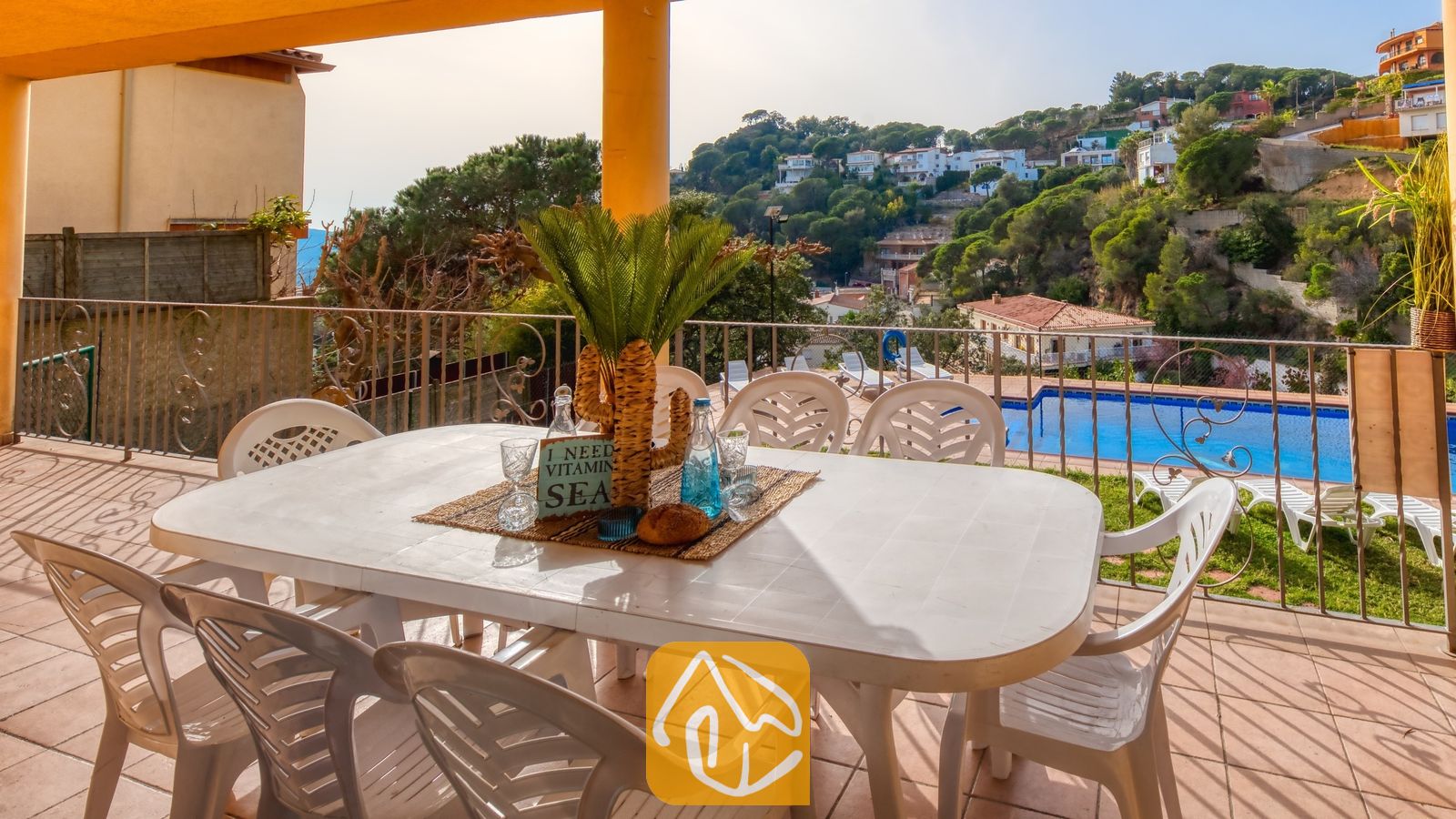 Vakantievilla's Costa Brava Spanje - Villa Marysol - Overdekt terras bij Villa Marysol met een witte kunststof eettafel, bijpassende stoelen en uitzicht op het zwembad tegen een achtergrond van heuvels.