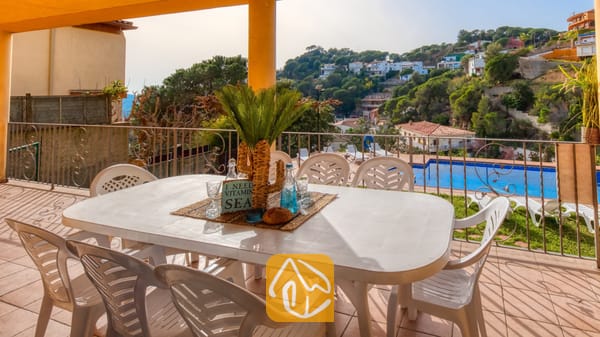 Vakantievilla's Costa Brava Spanje - Villa Marysol - Overdekt terras bij Villa Marysol met een witte kunststof eettafel, bijpassende stoelen en uitzicht op het zwembad tegen een achtergrond van heuvels.
