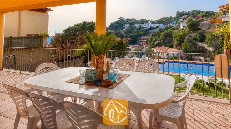 Vakantievilla's Costa Brava Spanje - Villa Marysol - Overdekt terras bij Villa Marysol met een witte kunststof eettafel, bijpassende stoelen en uitzicht op het zwembad tegen een achtergrond van heuvels.