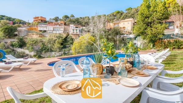 Vakantievilla's Costa Brava Spanje - Villa Marysol - Buiten eettafel met witte stoelen, waterflessen en servies naast een zwembad bij Villa Marysol, met ligstoelen en huizen op de heuvel op de achtergrond.