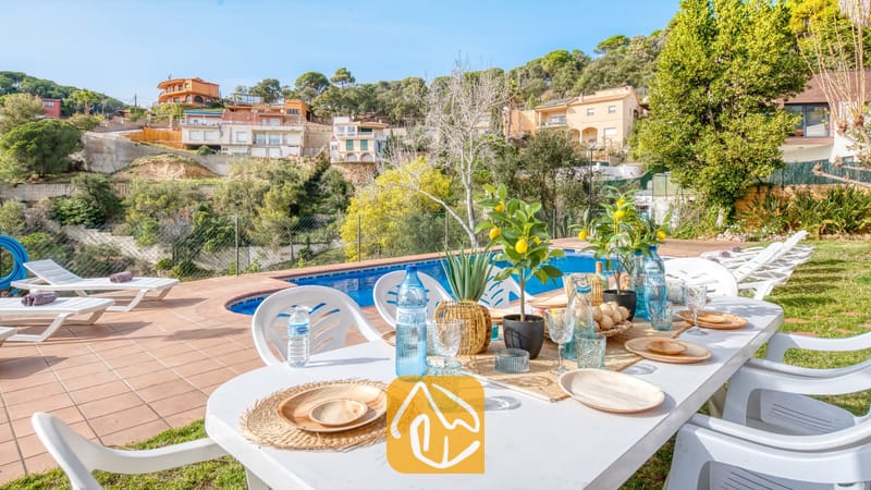 Vakantievilla's Costa Brava Spanje - Villa Marysol - Buiten eettafel met witte stoelen, waterflessen en servies naast een zwembad bij Villa Marysol, met ligstoelen en huizen op de heuvel op de achtergrond.