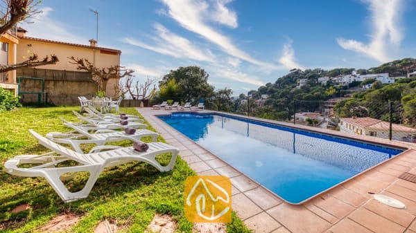 Vakantievilla's Costa Brava Spanje - Villa Marysol - Privézwembad met witte ligstoelen op een gazon en betegeld terras bij Villa Marysol, met uitzicht op de heuvels en het stadje onder een blauwe hemel.