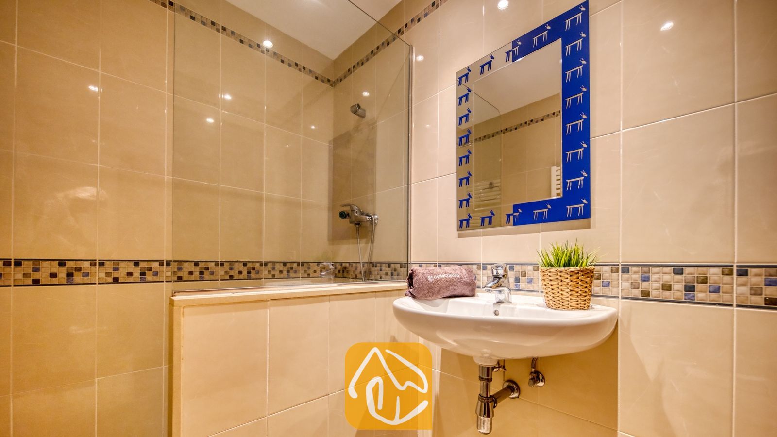 Vakantievilla's Costa Brava Spanje - Villa Marysol - Badkamer met betegelde muren in een beigetint, een glazen inloopdouche, een hangende witte wastafel en een blauwe spiegel met patroon.