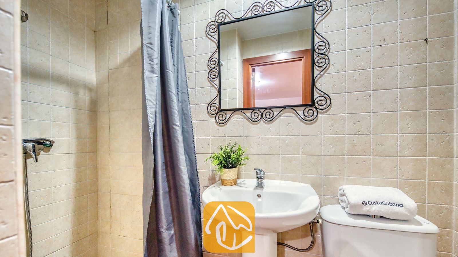 Vakantievilla's Costa Brava Spanje - Villa Marysol - Badkamer met beige betegelde muren, een vrijstaande wastafel, een toilet met een opgevouwen witte handdoek en een sierlijke spiegel met een zwarte lijst daarboven.