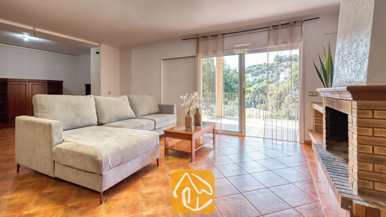 Vakantievilla's Costa Brava Spanje - Villa Marysol - Woonkamer van Villa Marysol met een grote grijze hoekbank, tegelvloeren, een stenen open haard en glazen schuifdeuren die opengaan naar een balkon met uitzicht op veel groen.