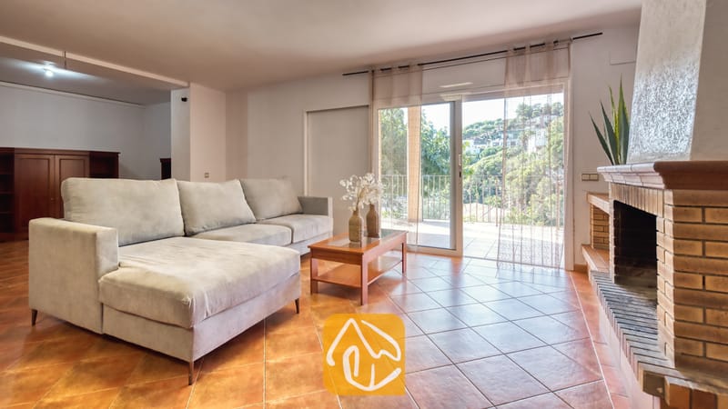 Vakantievilla's Costa Brava Spanje - Villa Marysol - Woonkamer van Villa Marysol met een grote grijze hoekbank, tegelvloeren, een stenen open haard en glazen schuifdeuren die opengaan naar een balkon met uitzicht op veel groen.