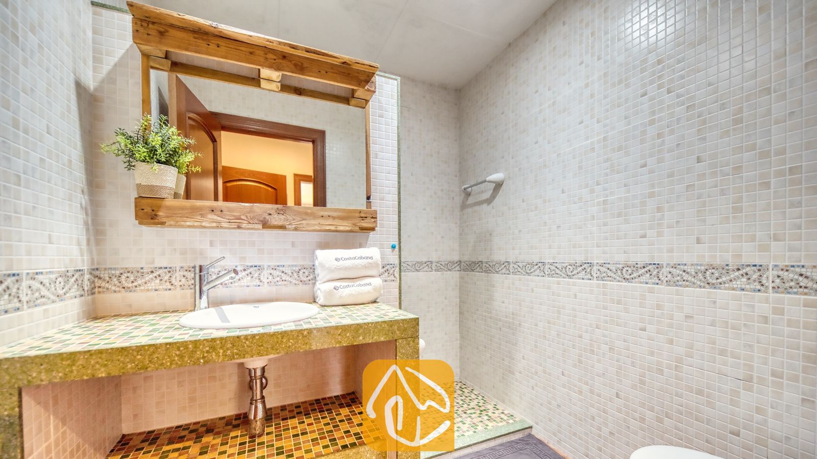 Vakantievilla's Costa Brava Spanje - Villa Marysol - Badkamer met een stenen wastafelmeubel voorzien van mozaïektegels, een houten spiegel met een hangplant en lichtbeige tegelmuren.
