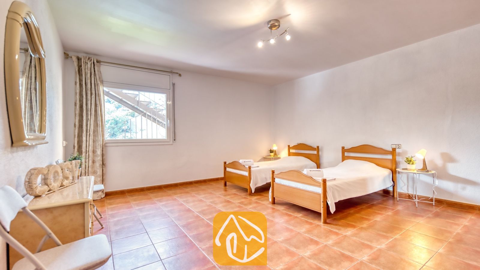 Vakantievilla's Costa Brava Spanje - Villa Marysol - Slaapkamer met twee houten eenpersoonsbedden, plavuizen vloeren, nachtkastlampjes en een raam met uitzicht op een buitentrap.