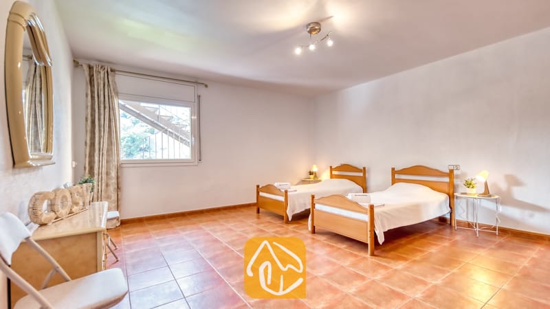 Vakantievilla's Costa Brava Spanje - Villa Marysol - Slaapkamer met twee houten eenpersoonsbedden, plavuizen vloeren, nachtkastlampjes en een raam met uitzicht op een buitentrap.