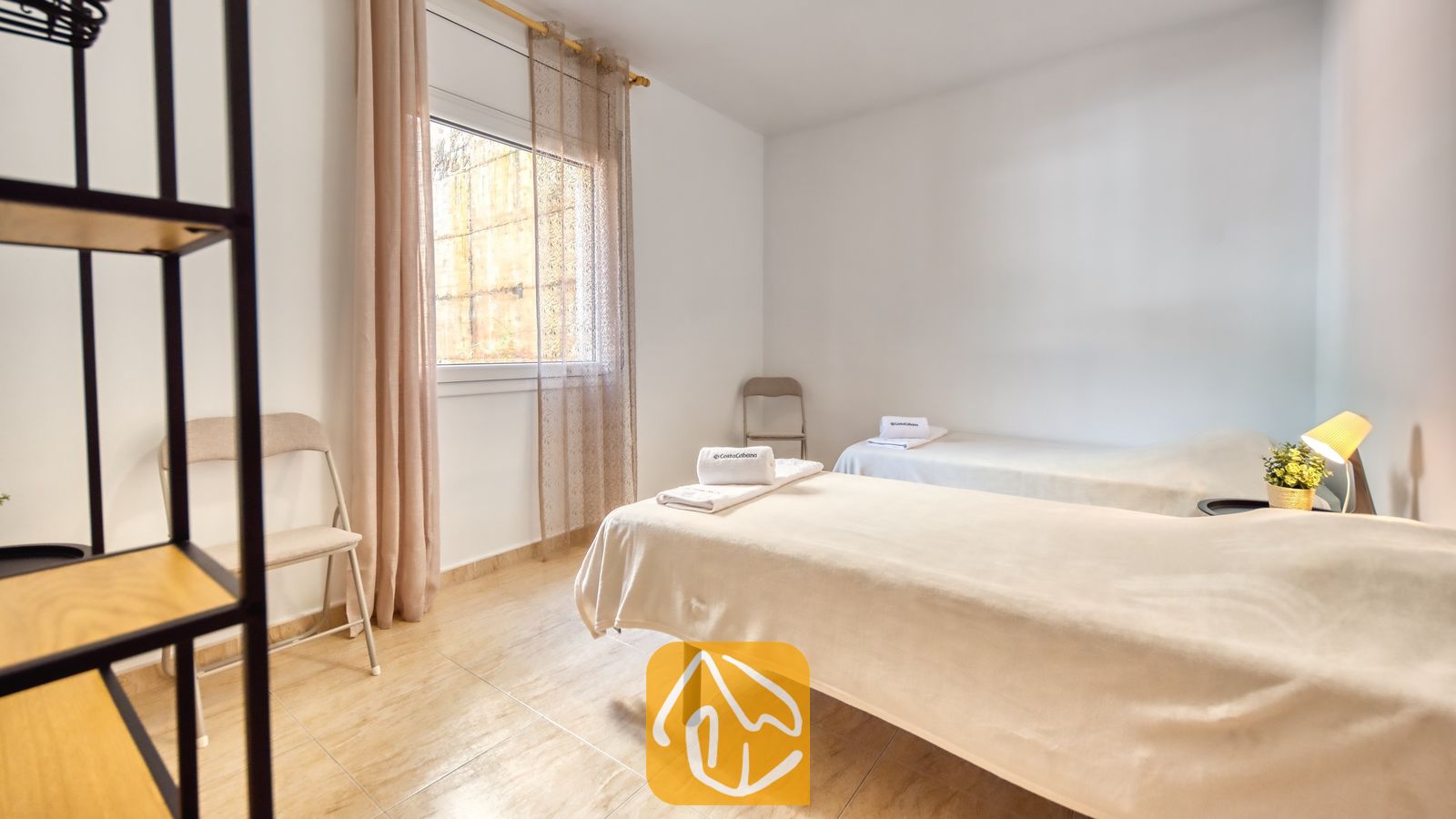 Vakantievilla's Costa Brava Spanje - Villa Marysol - Slaapkamer met twee eenpersoonsbedden, beddengoed in neutrale tinten, opgevouwen handdoeken, een klein nachtkastje en een raam met in-betweens.