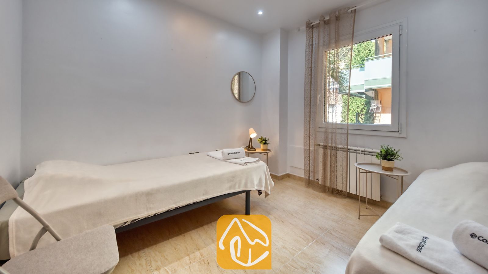 Vakantievilla's Costa Brava Spanje - Villa Marysol - Slaapkamer met twee eenpersoonsbedden opgemaakt met wit beddengoed, tegelvloer, een ronde wandspiegel en een raam met inbetweens.