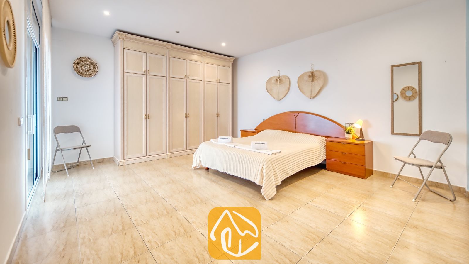 Vakantievilla's Costa Brava Spanje - Villa Marysol - Slaapkamer met een tweepersoonsbed, houten hoofdbord, grote witte kledingkast, tegelvloer en twee klapstoelen.