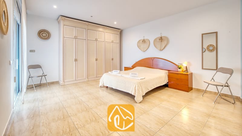 Vakantievilla's Costa Brava Spanje - Villa Marysol - Slaapkamer met een tweepersoonsbed, houten hoofdbord, grote witte kledingkast, tegelvloer en twee klapstoelen.