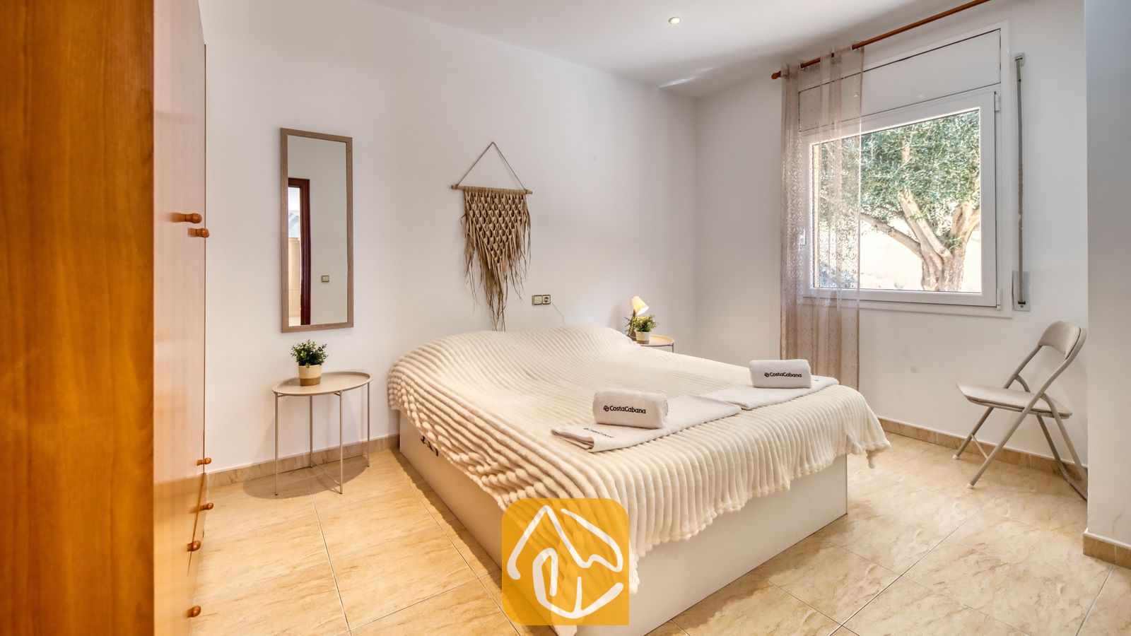 Vakantievilla's Costa Brava Spanje - Villa Marysol - Slaapkamer met een tweepersoonsbed voorzien van een witte, textuurrijke sprei, opgevouwen handdoeken, een rechthoekige wandspiegel en een klein raam met uitzicht op groene bomen.