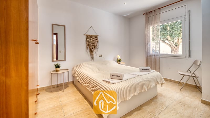 Vakantievilla's Costa Brava Spanje - Villa Marysol - Slaapkamer met een tweepersoonsbed voorzien van een witte, textuurrijke sprei, opgevouwen handdoeken, een rechthoekige wandspiegel en een klein raam met uitzicht op groene bomen.