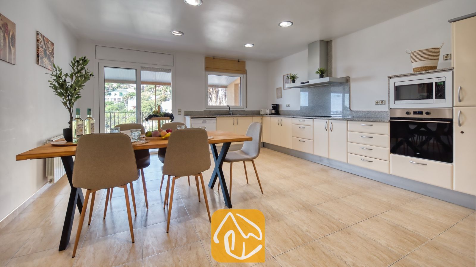 Vakantievilla's Costa Brava Spanje - Villa Marysol - Open keuken en eethoek met een houten tafel, beige gestoffeerde stoelen, lichte kasten en een schuifpui met uitzicht op het groen.