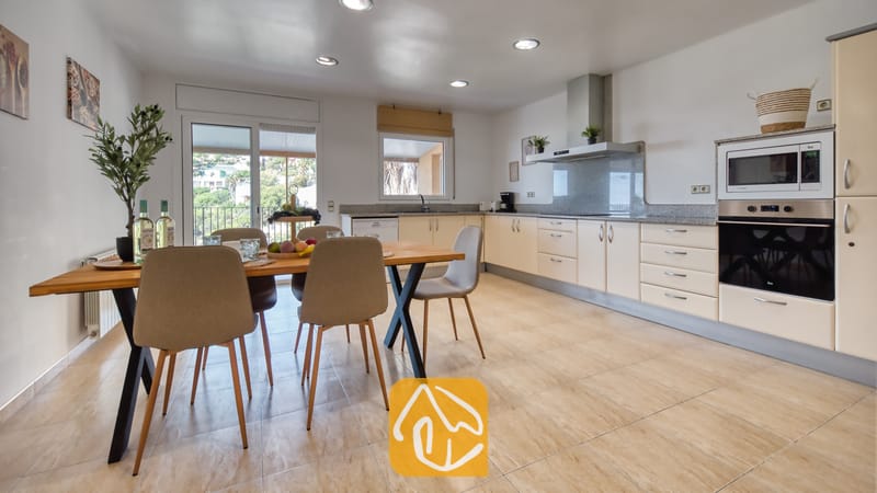 Vakantievilla's Costa Brava Spanje - Villa Marysol - Open keuken en eethoek met een houten tafel, beige gestoffeerde stoelen, lichte kasten en een schuifpui met uitzicht op het groen.