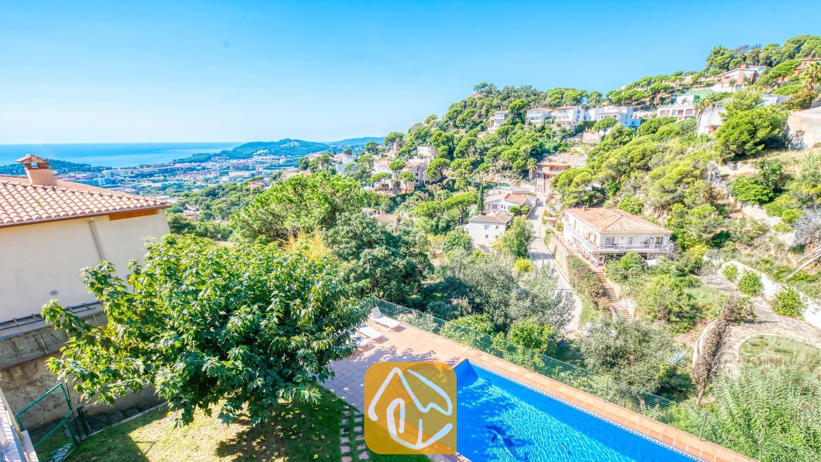Vakantievilla's Costa Brava Spanje - Villa Marysol - Verheven uitzicht vanaf Villa Marysol op een blauw zwembad op een betegeld terras, groene bomen en huizen die langs een heuvel afdalen richting de zee onder een heldere hemel.