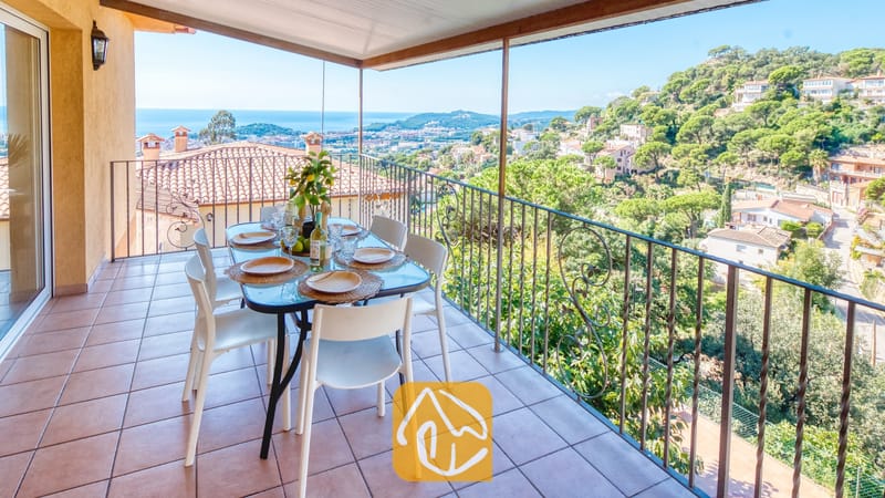 Vakantievilla's Costa Brava Spanje - Villa Marysol - Overdekt terras bij Villa Marysol met een eettafel, witte stoelen en een metalen balustrade met uitzicht op een stad en de zee.