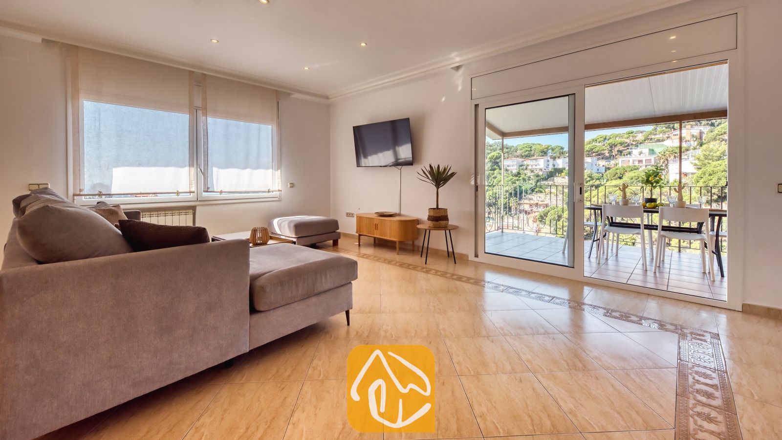 Vakantievilla's Costa Brava Spanje - Villa Marysol - Woonkamer van Villa Marysol, voorzien van een grijze hoekbank, een flatscreentelevisie aan de muur, tegelvloeren en glazen schuifdeuren die opengaan naar een gemeubileerd balkon.