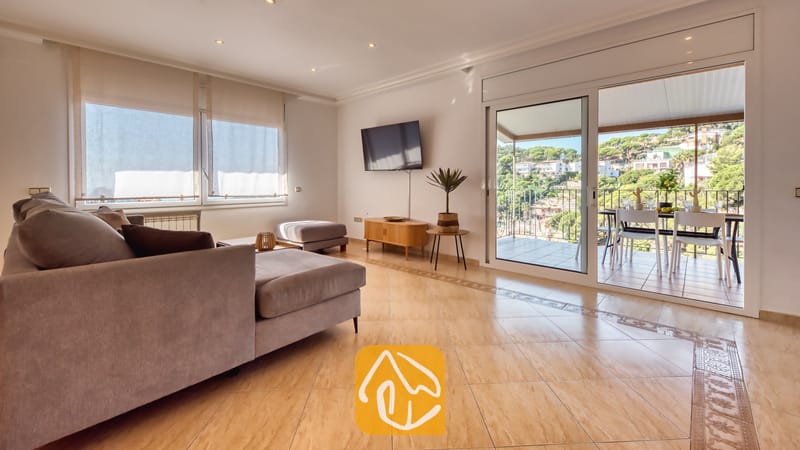 Vakantievilla's Costa Brava Spanje - Villa Marysol - Woonkamer van Villa Marysol, voorzien van een grijze hoekbank, een flatscreentelevisie aan de muur, tegelvloeren en glazen schuifdeuren die opengaan naar een gemeubileerd balkon.