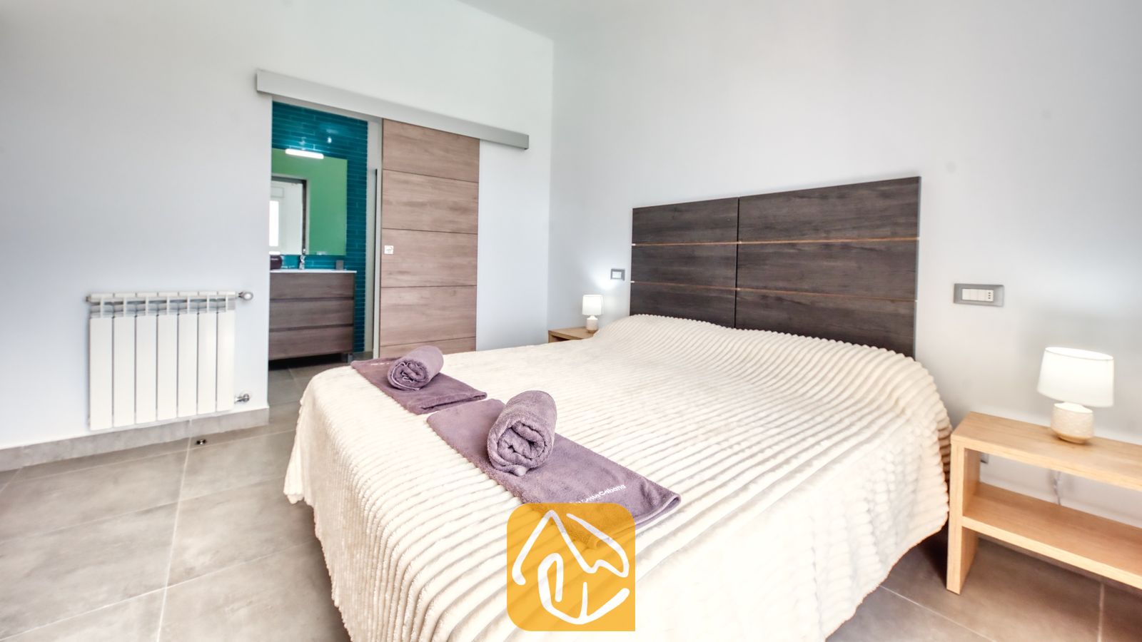 Vakantievilla's Costa Brava Spanje - Villa BellaVista - Slaapkamer in Villa BellaVista, voorzien van een groot bed met een donkerhouten hoofdbord, paarse handdoeken en een schuifdeur die leidt naar een en-suite badkamer met blauwe tegels.