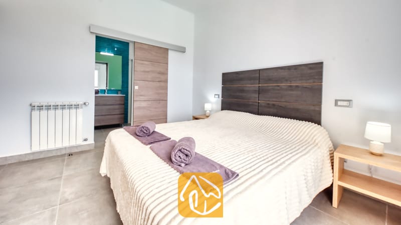 Vakantievilla's Costa Brava Spanje - Villa BellaVista - Slaapkamer in Villa BellaVista, voorzien van een groot bed met een donkerhouten hoofdbord, paarse handdoeken en een schuifdeur die leidt naar een en-suite badkamer met blauwe tegels.
