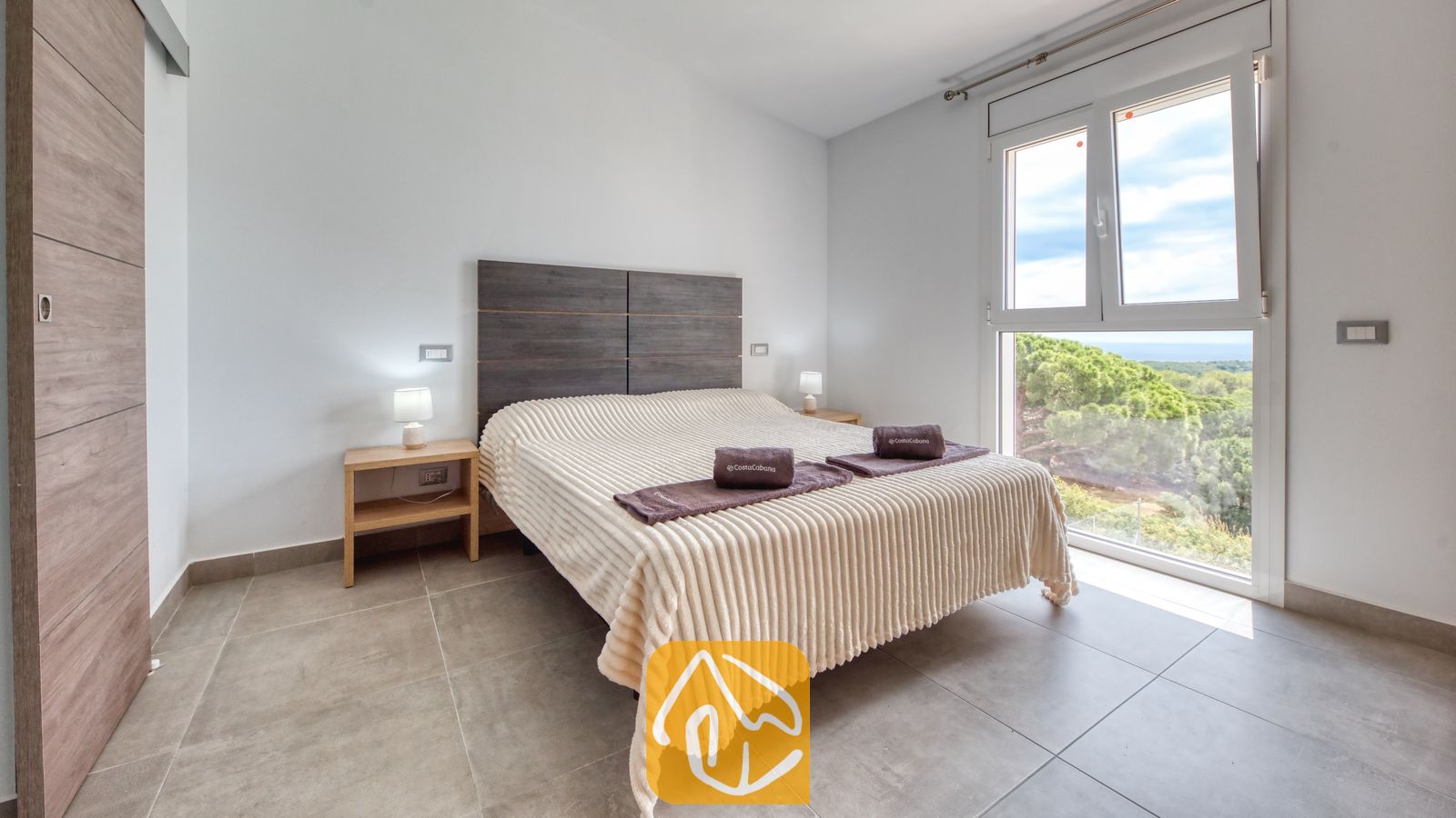 Vakantievilla's Costa Brava Spanje - Villa BellaVista - Slaapkamer met een crèmekleurige, geribbelde sprei, een donkerhouten hoofdbord en nachtkastlampen. Een groot raam biedt uitzicht op een groen boslandschap onder een blauwe lucht.