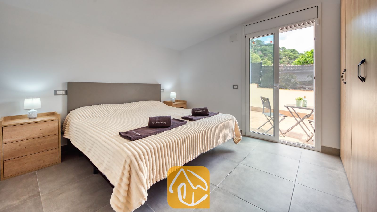 Vakantievilla's Costa Brava Spanje - Villa BellaVista - Slaapkamer in Villa BellaVista, voorzien van een tweepersoonsbed met een geribbelde crèmekleurige plaid, houten nachtkastjes met bijpassende lampen en een glazen deur die uitkomt op een terras met zitgelegenheid.