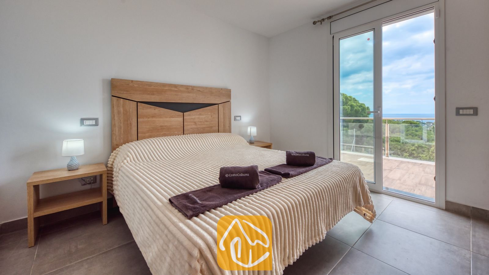 Vakantievilla's Costa Brava Spanje - Villa BellaVista - Slaapkamer in Villa BellaVista met een crèmekleurige sprei met textuur, een houten hoofdbord en glazen schuifdeuren die uitkomen op een balkon met uitzicht op zee.