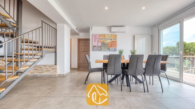 Vakantievilla's Costa Brava Spanje - Villa BellaVista - Eethoek in Villa BellaVista met een houten tafel en grijze stoelen, een moderne trap met metalen leuningen en grote glazen deuren die uitkomen op een terras.