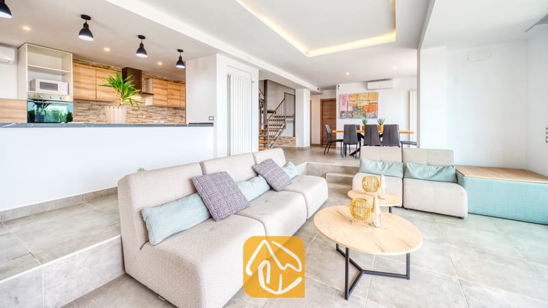 Vakantievilla's Costa Brava Spanje - Villa BellaVista - Het woongedeelte van Villa BellaVista is voorzien van een beige modulaire bank met pastelkleurige kussens, ronde houten salontafels en een moderne keuken met lichthouten kasten.