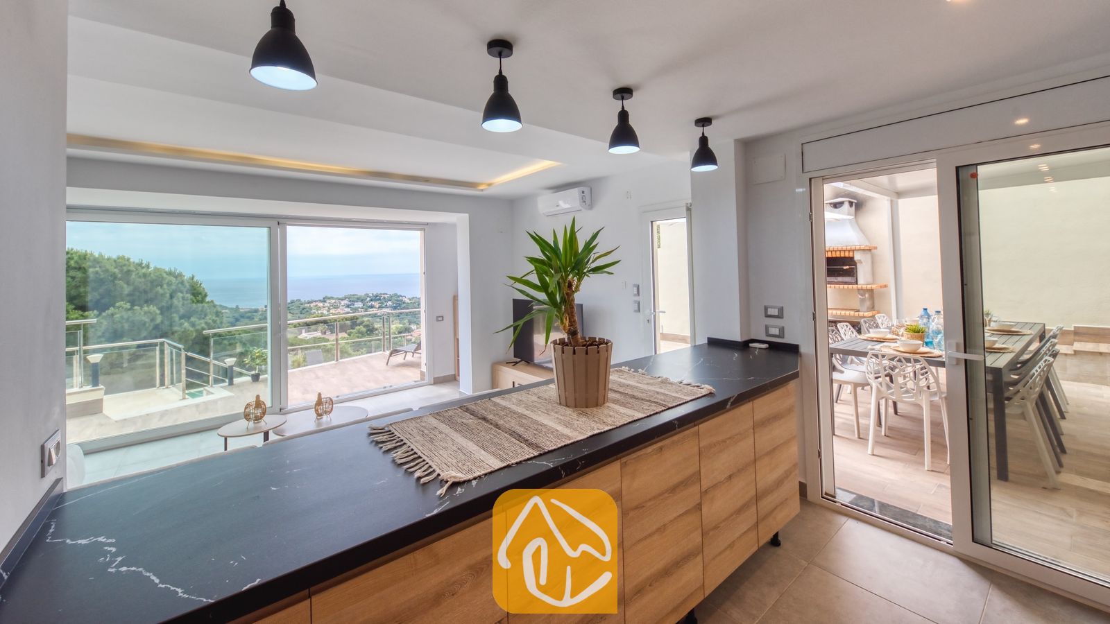 Vakantievilla's Costa Brava Spanje - Villa BellaVista - Kookeiland met een werkblad in zwart marmerlook en houten kasten in Villa BellaVista. Zwarte hanglampen sieren het plafond en bieden uitzicht op het terras met zeezicht door de grote raampartijen.