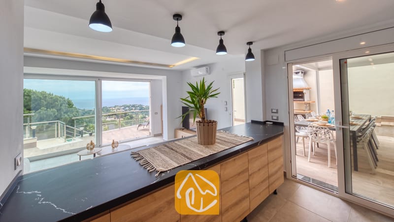 Vakantievilla's Costa Brava Spanje - Villa BellaVista - Kookeiland met een werkblad in zwart marmerlook en houten kasten in Villa BellaVista. Zwarte hanglampen sieren het plafond en bieden uitzicht op het terras met zeezicht door de grote raampartijen.