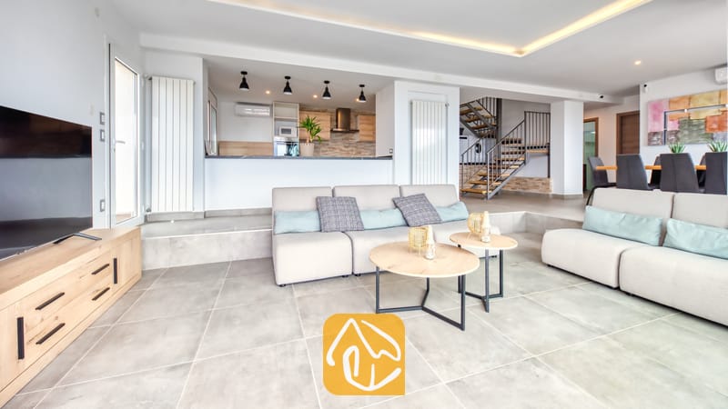 Vakantievilla's Costa Brava Spanje - Villa BellaVista - De open woonruimte van Villa BellaVista is voorzien van beige banken met blauwe kussens, ronde houten salontafels en een tegelvloer die overloopt in een keuken en een metalen trap.