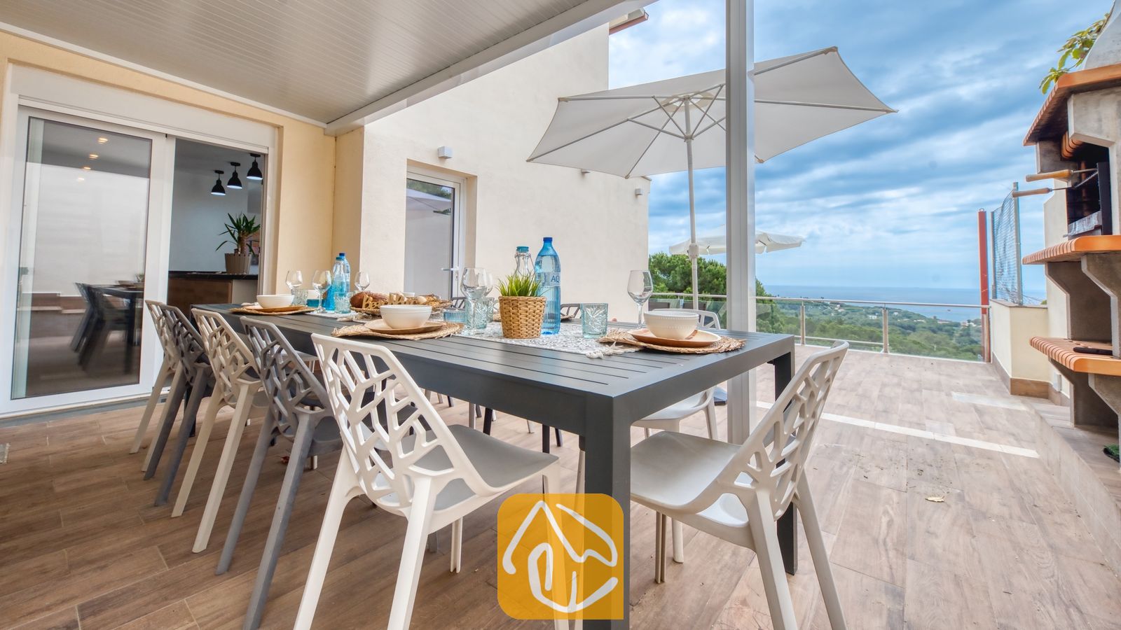 Vakantievilla's Costa Brava Spanje - Villa BellaVista - Terras van Villa BellaVista met een zwarte eettafel, witte geometrische stoelen, een ingebouwde barbecue en uitzicht op de verre kustlijn.
